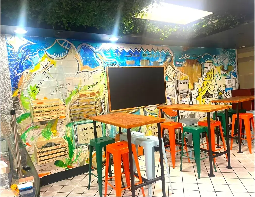A restaurant wall painted as a Jerusalem market, with Hebrew street names and a menorah, above orange and green stools