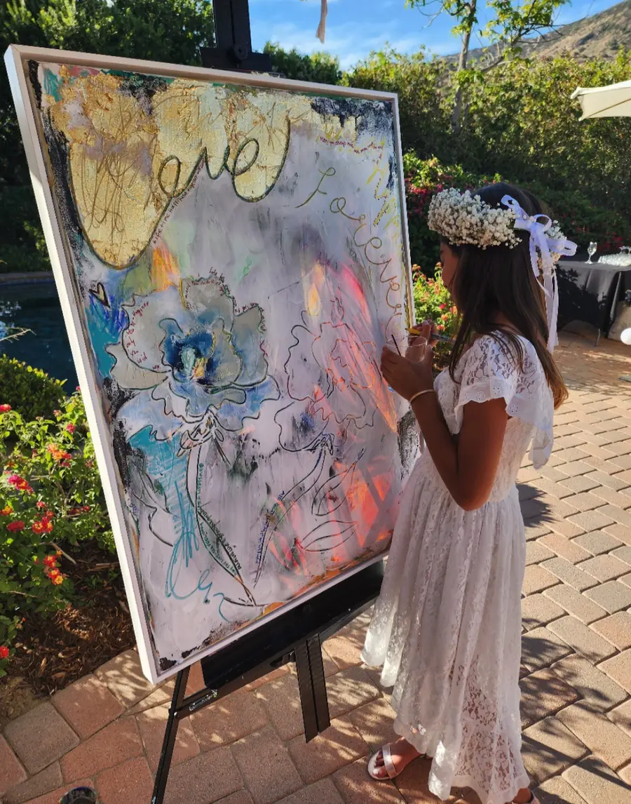 A young guest in a white dress and flower crown paints on a gold and blue wedding canvas